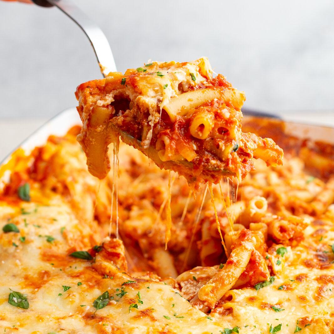 baked ziti in a casserole dish with a hand using a scoop to pull some of the baked ziti out of the pan