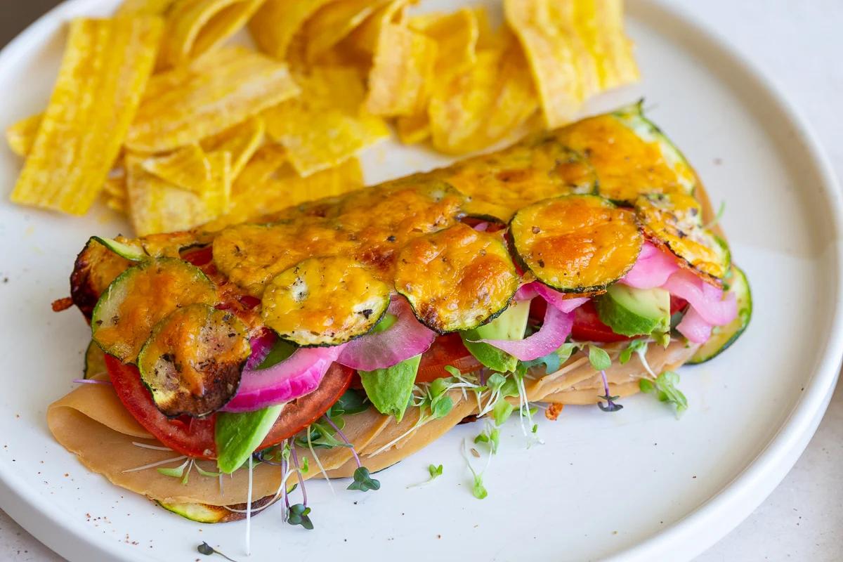 cheesy zucchini wrap stuffed with vegan meat, red onion, avocado and tomato. The wrap is on a plate with chips