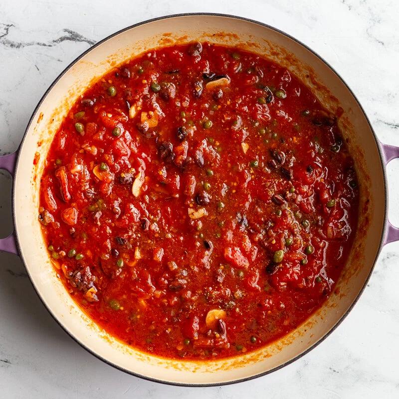 Puttanesca tomato sauce simmering in a skillet with olives and capers.