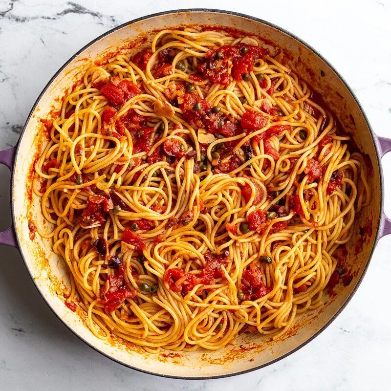 Spaghetti puttanesca tossed together in a skillet with sauce, olives, and capers.