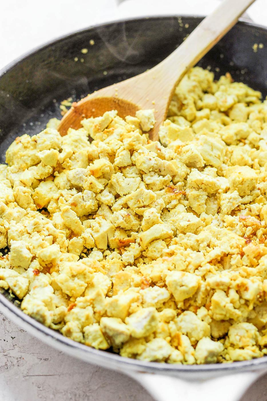 Scrambling tofu in a skillet with a wooden spoon.