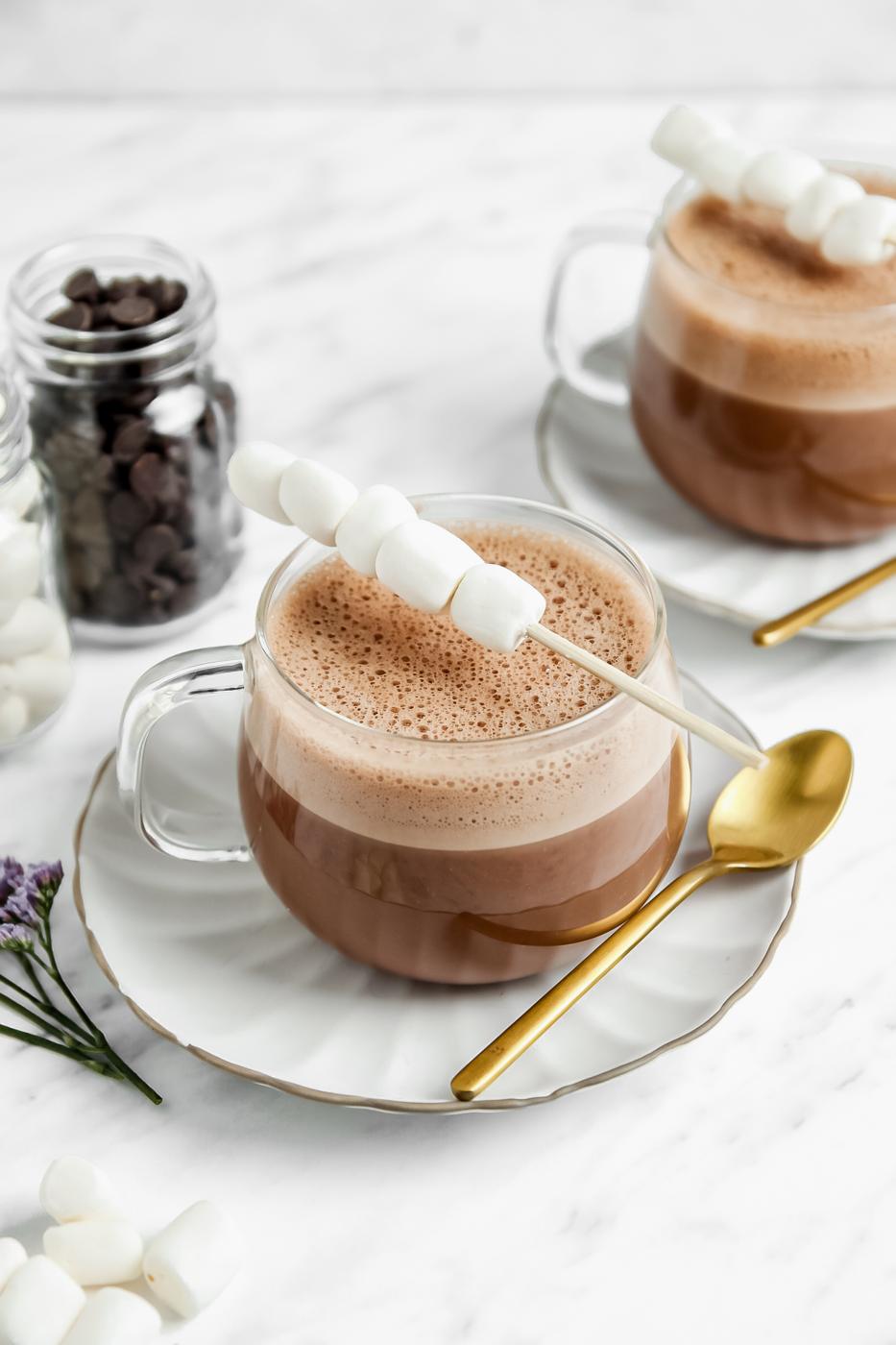 vegan hot chocolate in a clear glass with a stick of marshmallows on top of them. more hot chocolate in the background