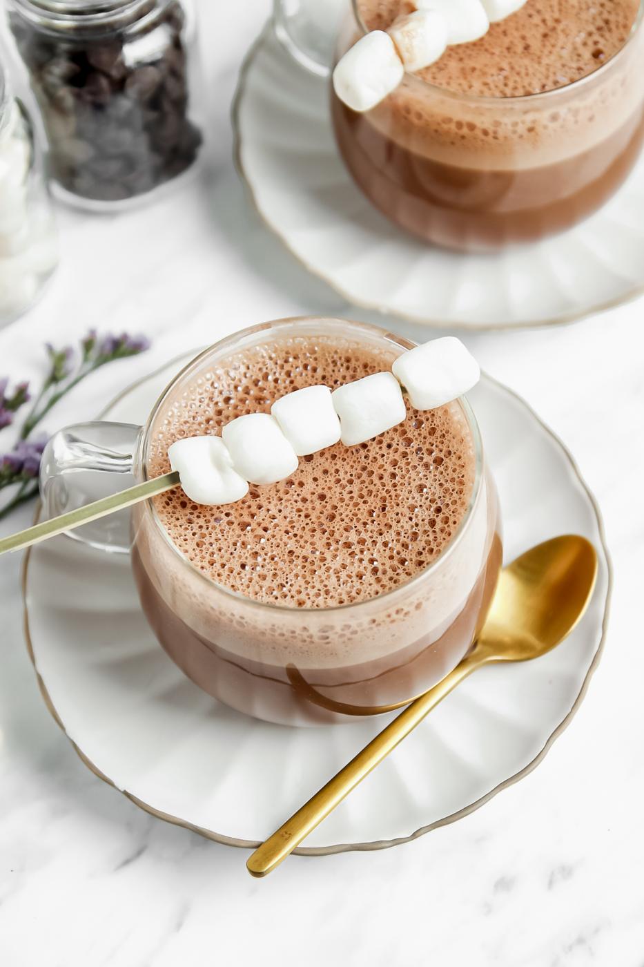 vegan hot chocolate in a clear glass with a stick of marshmallows on top of them. more hot chocolate in the background