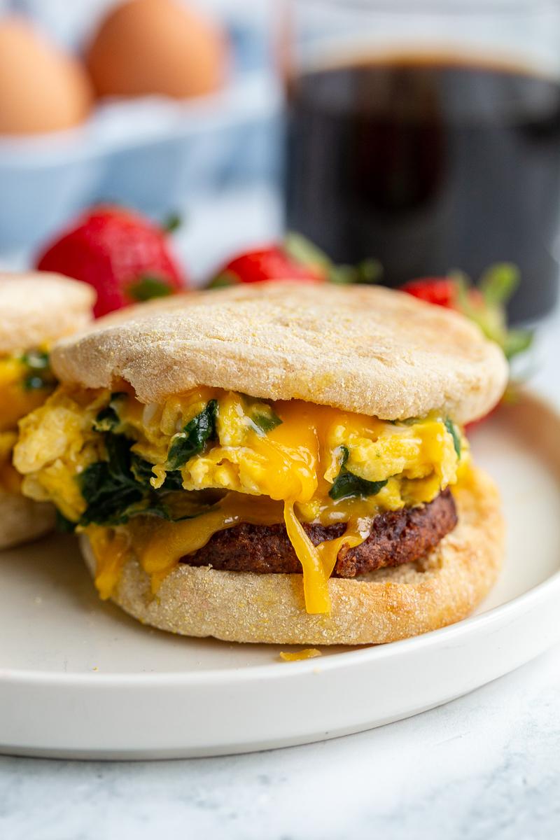 english muffin sandwich with spinach scrambled eggs and sausage topped with cheese in the middle. strawberries and coffee are in the background