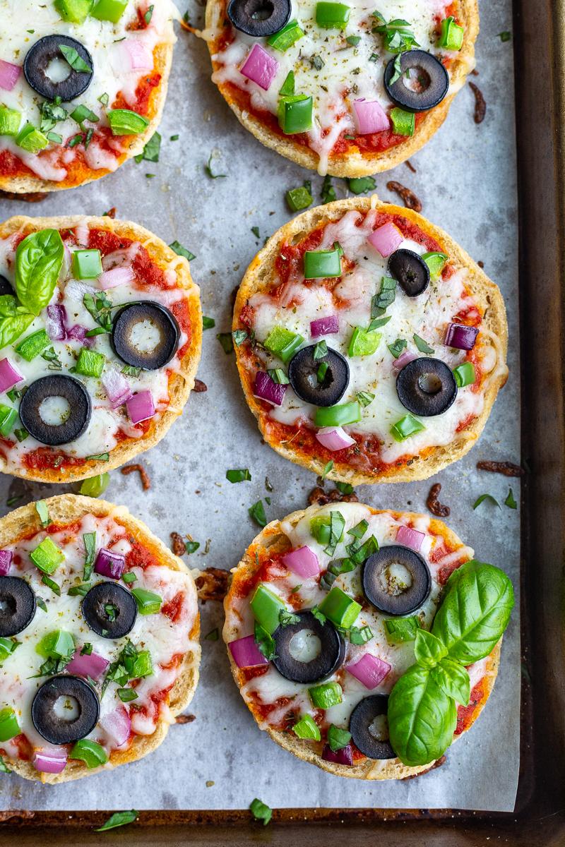 close up shot of english muffin pizzas (topped with cheese, red onion, green pepper, olives, and basil) sitting on a parchment lined baking sheet