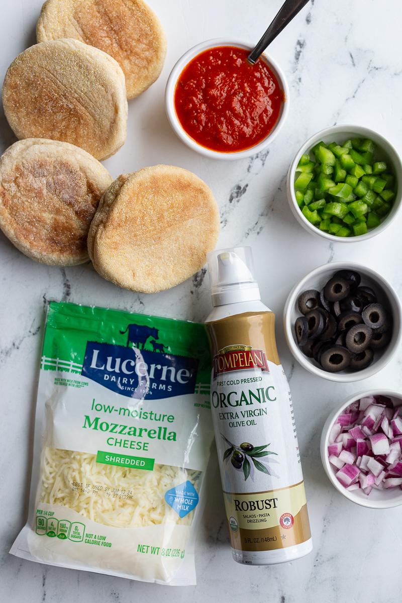 ingredients layed out for english muffin pizzas: english muffins, cooking spray, mozzarella cheese, diced onions, olives, and bell pepper, pizza sauce.