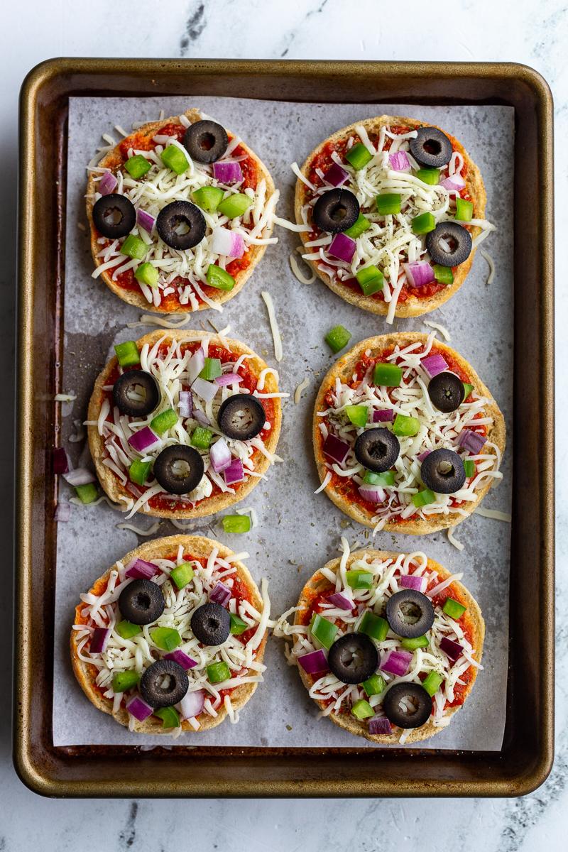 english muffin pizzas (topped with cheese, red onion, green pepper, olives, and basil) sitting on a parchment lined baking sheet before they've been baked