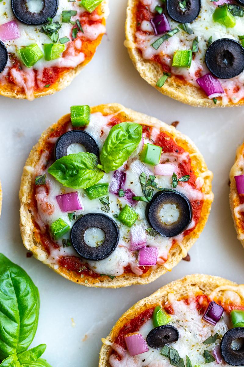 close up shot of english muffin pizzas (topped with cheese, red onion, green pepper, olives, and basil) sitting on a white board