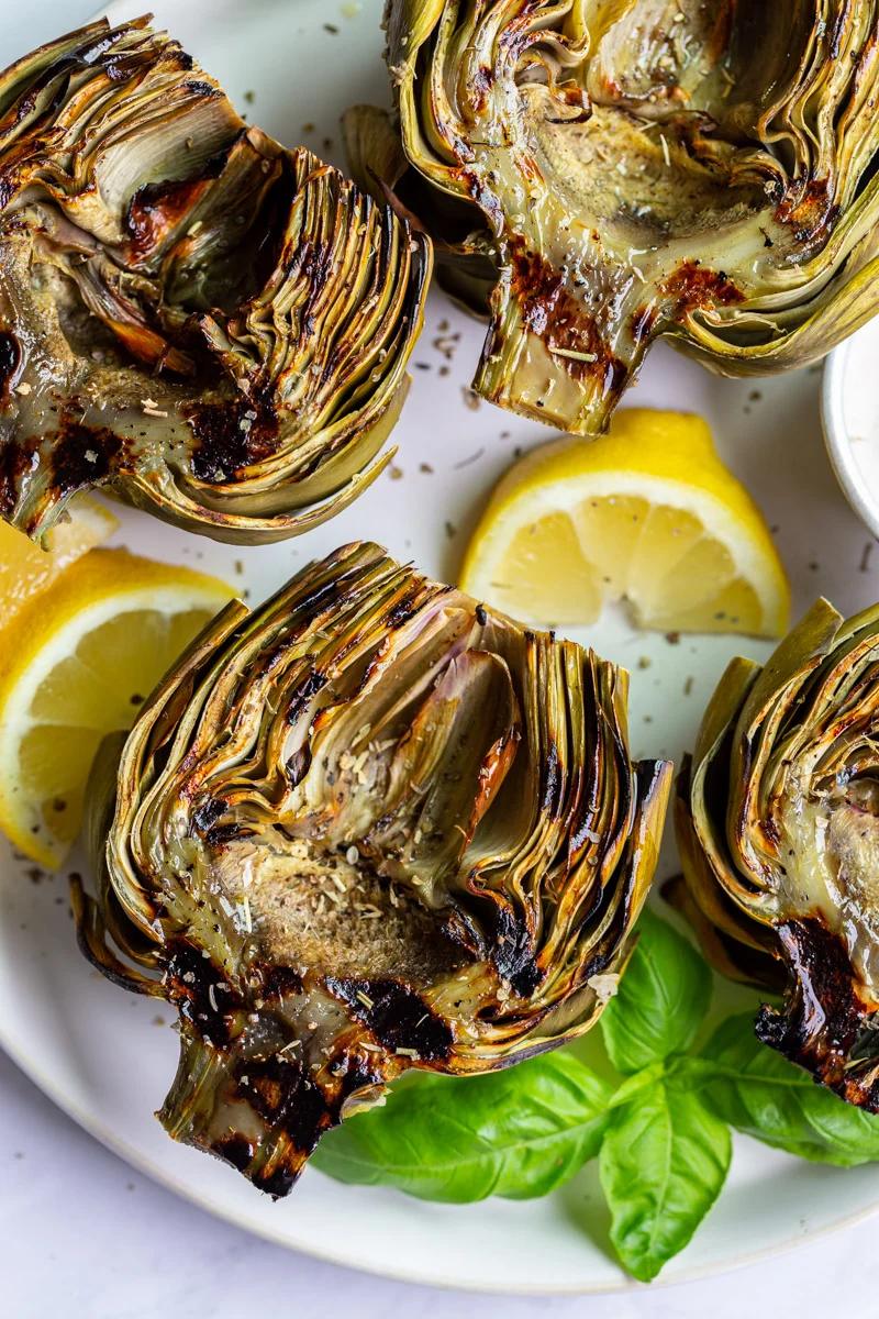 large plate with 4 grilled artichokes on it and lemon slices on the plate