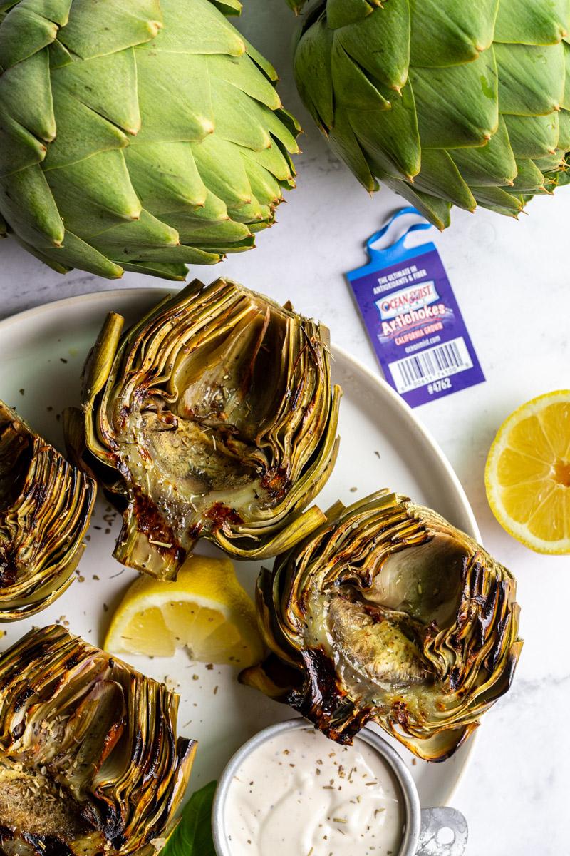 large plate with 4 grilled artichokes on it and lemon slices on the plate. fresh artichokes in the corner