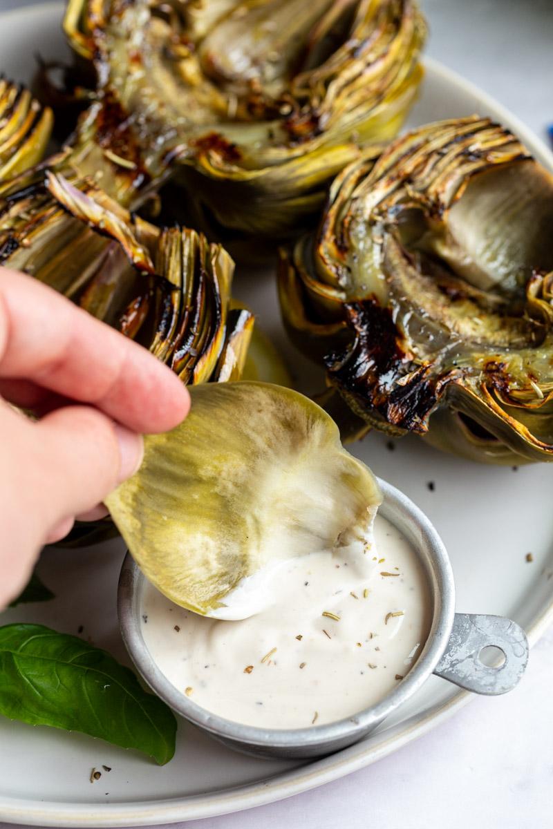 large plate with 4 grilled artichokes on it and lemon slices on the plate. hand holding an artichoke leaf in dip
