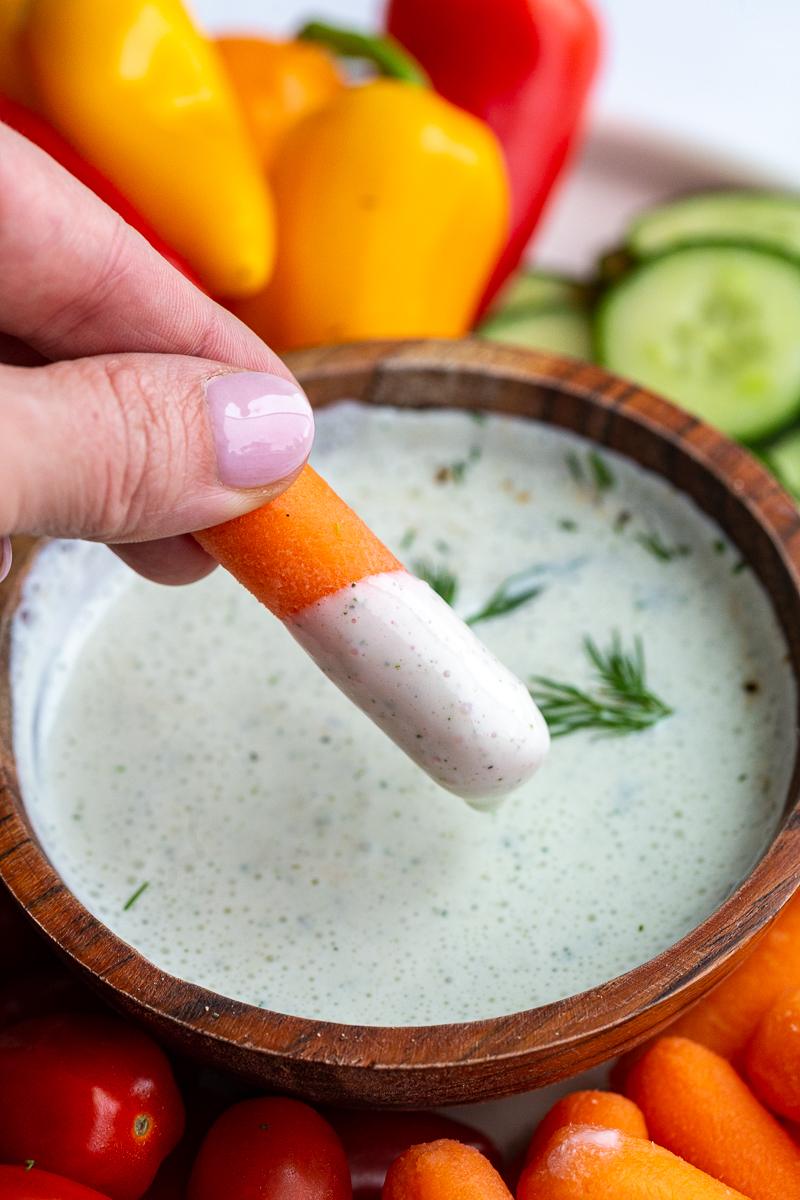 cottage cheese ranch in a wood bowl topped with fresh dill. The bowl is surrounded by fresh colorful veggies and crackers. There's a hand holding a carrot stick with ranch on it