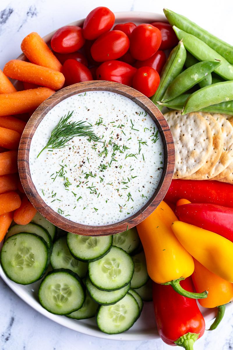 cottage cheese ranch in a wood bowl topped with fresh dill. The bowl is surrounded by fresh colorful veggies and crackers