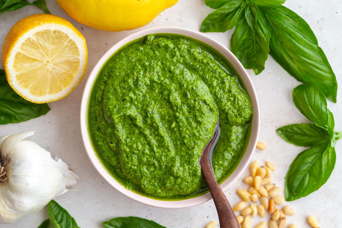 from-scratch pesto recipe - a bowl of pesto with a wooden spoon in it and lemons and fresh basil in the corners of the shot
