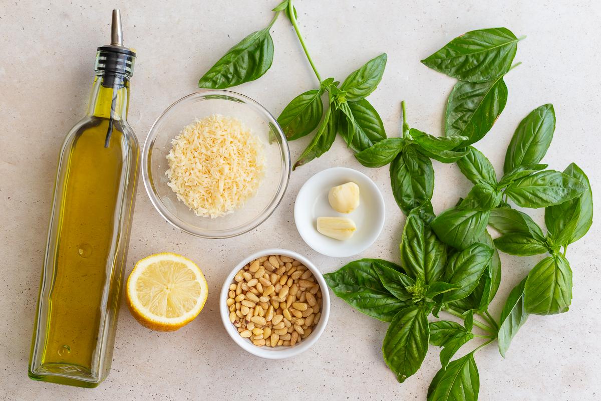 ingredients laid out to make from-scratch pesto including olive oil, parmesan, toasted pinenuts, fresh basil, lemon, and garlic
