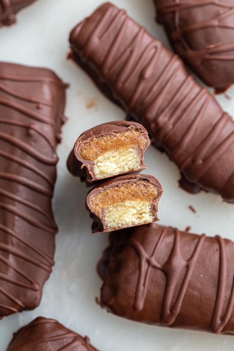 top down shot of from-scratch twix chocolate bars. There's extra chocolate drizzled on top of them in a swirled pattern. One or the bars is cut in half so that the can see the caramel and shortbread fiilling