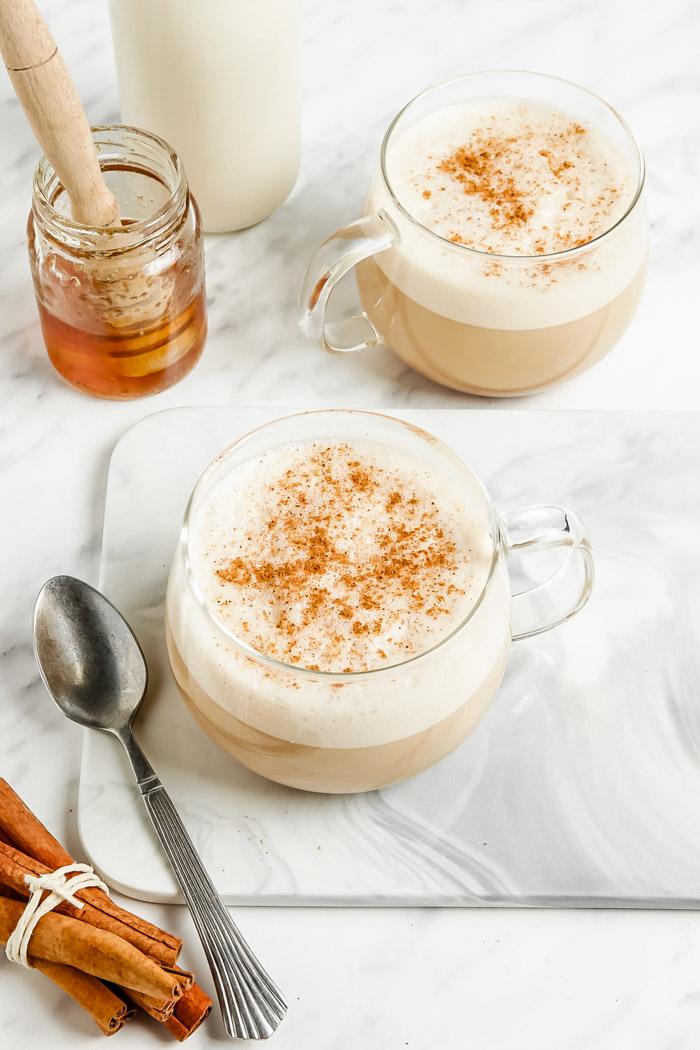 oat milk lattes in a clear glass with lots of foam and cinnamon on top. Honey and milk in the background of the shot and fresh cinnamon sticks in the front