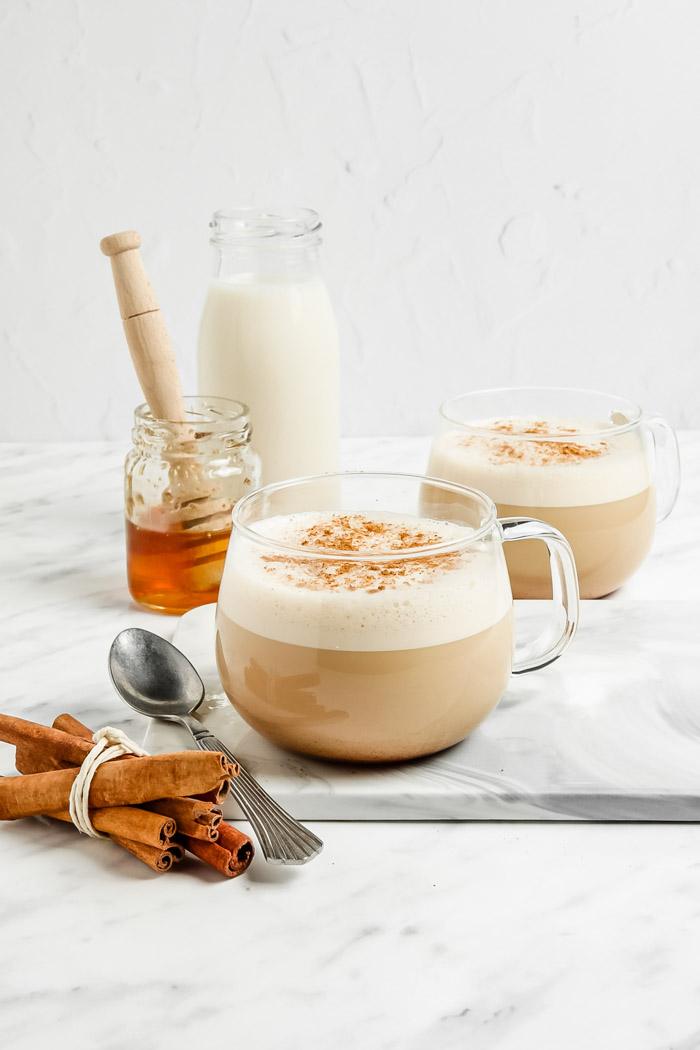 oat milk lattes in a clear glass with lots of foam and cinnamon on top. Honey and milk in the background of the shot and fresh cinnamon sticks in the front