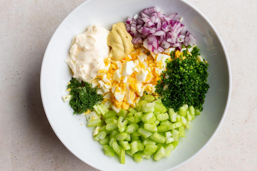 ingredients in large bowl to make from-scratch egg salad including hard boiled eggs chopped up, red onion, chives, mayo, and celery