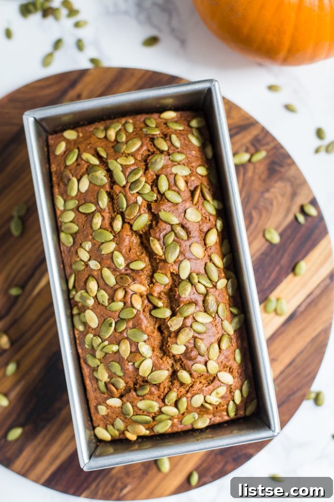 Plant-Based Pumpkin Spice Loaf 3 Top-down view of a golden brown vegan pumpkin bread loaf in a pan, generously topped with roasted pepitas.