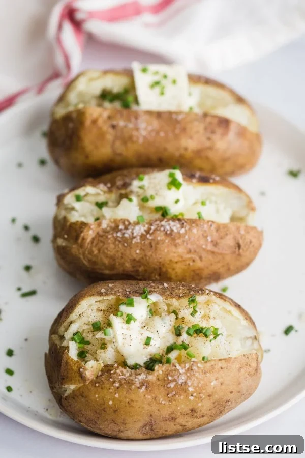 Fluffy Instant Pot Baked Potatoes 6 Close-up of a large plate filled with several Instant Pot baked potatoes, each split open and adorned with a dollop of butter and a scattering of fresh chives. A textured red and white kitchen towel rests in the background, completing the inviting culinary scene.