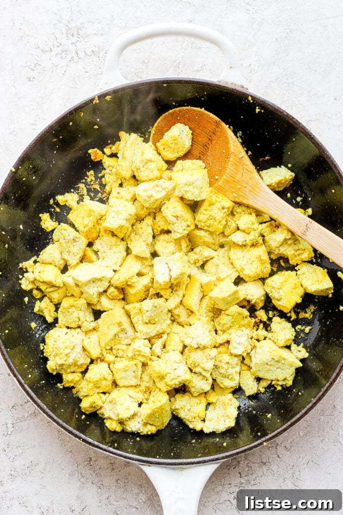 Effortless Vegan Scramble 5 A wooden spoon crumbling large tofu chunks in a hot non-stick pan, initiating the tofu scramble cooking process.