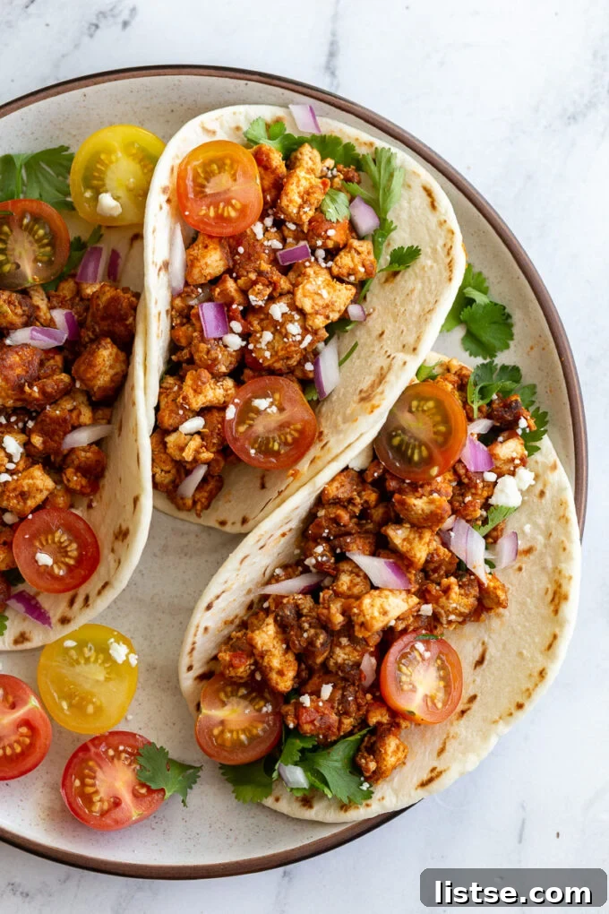 Effortless Tofu Taco Bliss 5 Three soft flour tortillas generously filled with savory tofu taco meat, and vibrant toppings including halved cherry tomatoes, fresh cilantro leaves, finely chopped red onion, and crumbled vegan feta cheese.