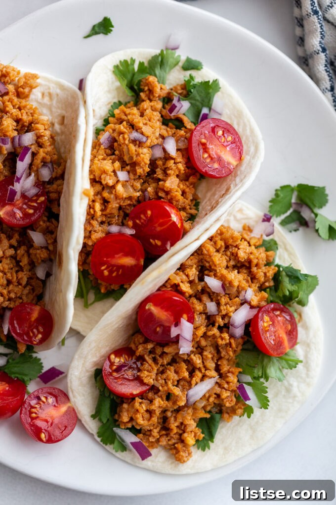 Three tacos on a plate topped with cilantro, TVP taco meat, cherry tomatoes, and onion, showcasing a delicious vegan meal.