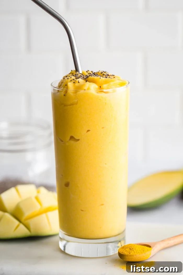 Tall glass of Golden Milk Mango Smoothie with turmeric, garnished with chia seeds. Fresh mango and a spoon of turmeric are visible in the soft-focus background.