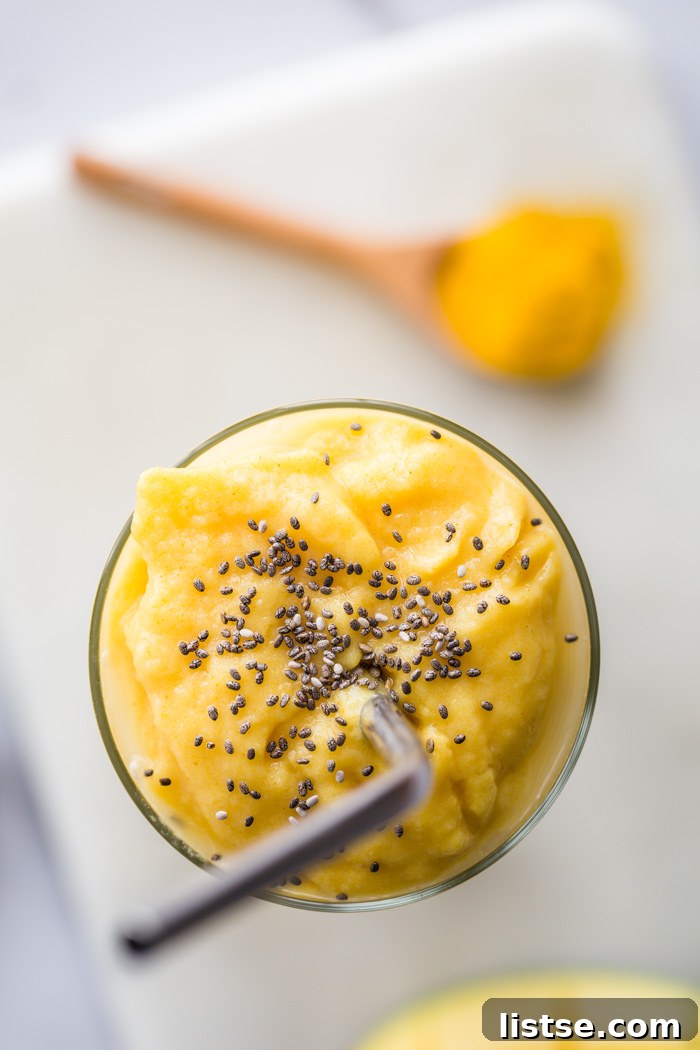 Golden Milk Mango Smoothie in a tall glass, with whole mango and a spoon of turmeric on the side. The smoothie is topped with a sprinkle of chia seeds.