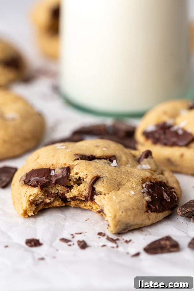 Decadent Vegan Chocolate Chip Cookies 4 chocolate chip cookie with a bit taken out of it. More cookies, chocolate chips, and glass of milk in the background