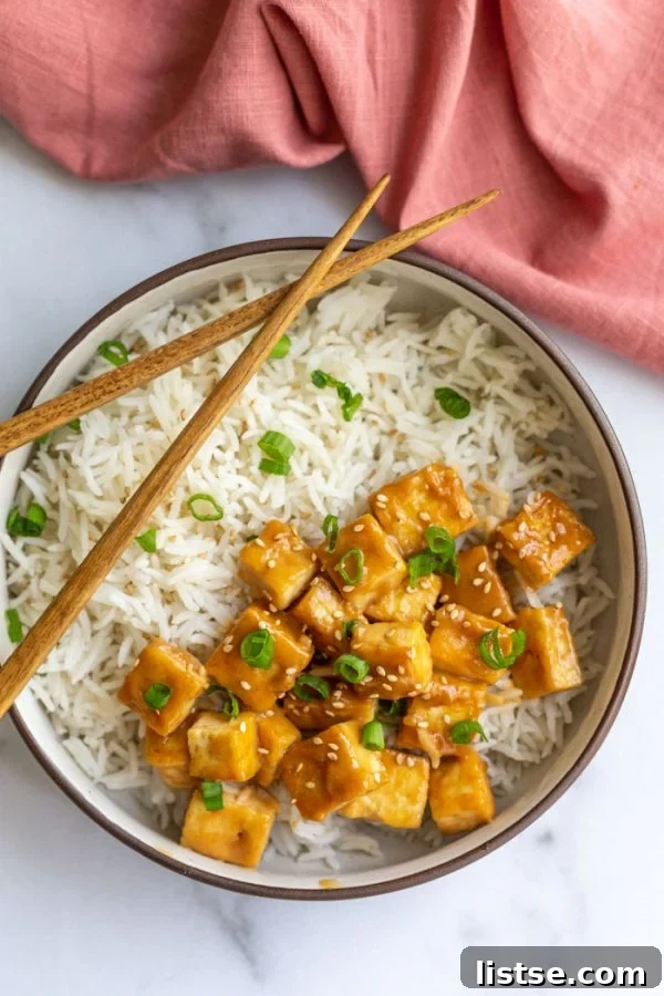 sauteed tofu on a bed of rice and topped with green onion and sesame seeds. on a marble board with a pink towel in the corner and chopsticks resting on the side of the bowl