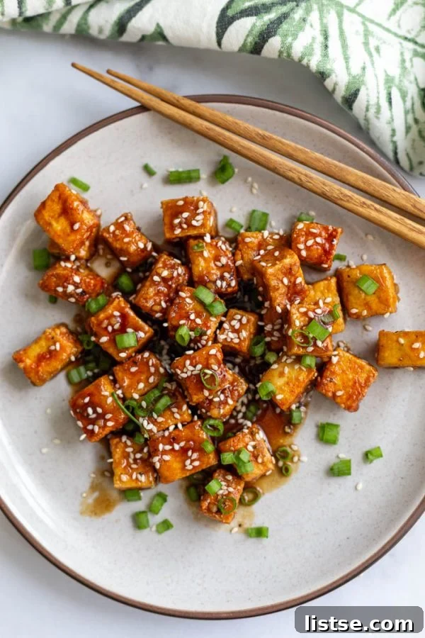 Top-down shot of crispy air fryer tofu cubes on a plate, coated in teriyaki sauce and garnished with green onion and sesame seeds, with chopsticks nearby.