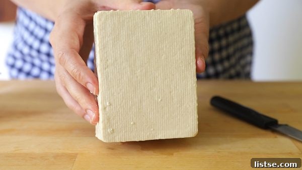 Hands holding a block of firm tofu over a butcher block with a knife in the background, illustrating raw tofu.