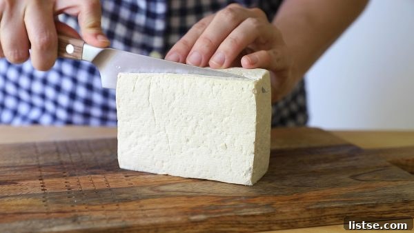 Hands cutting a block of tofu in half on a butcher block counter, preparing it for cooking.