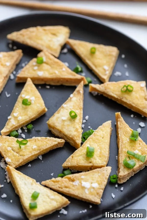 Side shot of perfectly pan-fried tofu triangles, lightly golden and crispy, garnished with flaky sea salt and green onions on a white plate.