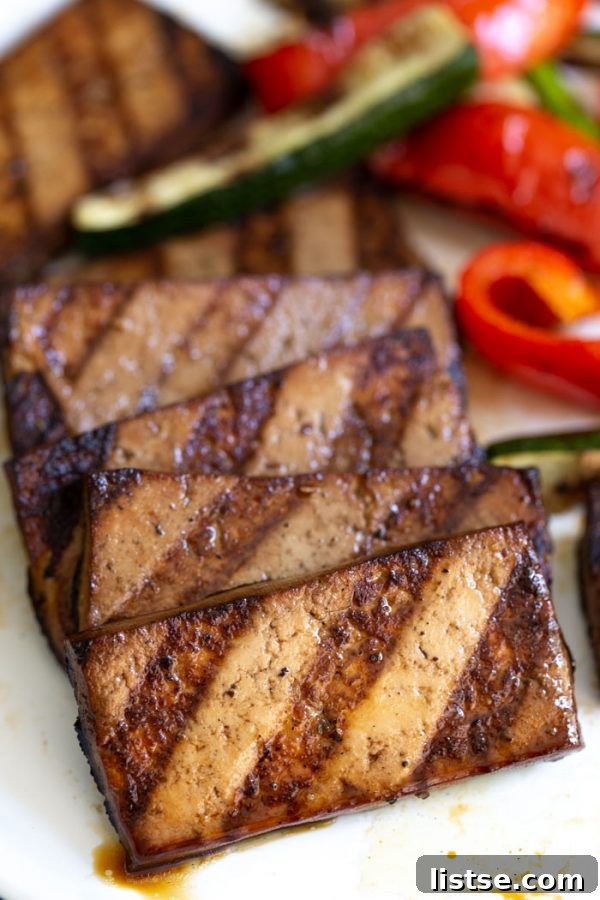 Side shot of marinated grilled tofu with prominent grill marks, accompanied by grilled red bell pepper and zucchini slices in the background.