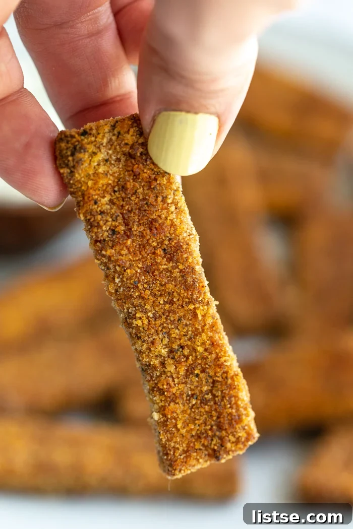 Up close shot of a hand holding a perfectly golden-brown tofu fish stick, showing its crispy breading.