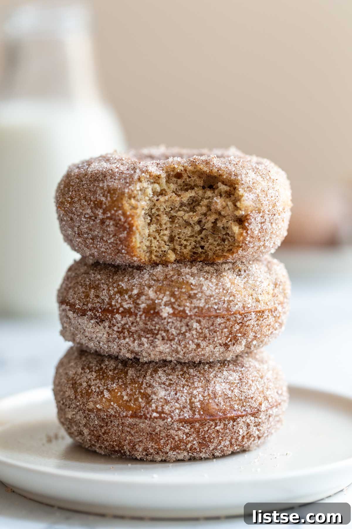 Three golden-brown baked vegan donuts stacked with one having a bite taken out, covered in cinnamon sugar, on a wooden board.