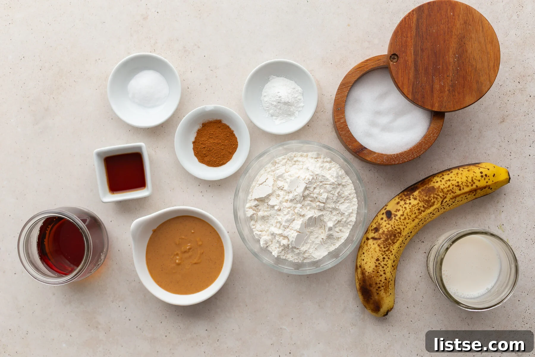 Fresh ingredients laid out on a marble surface, including bananas, maple syrup, flour, nut butter, and almond milk, ready for making vegan banana donuts.