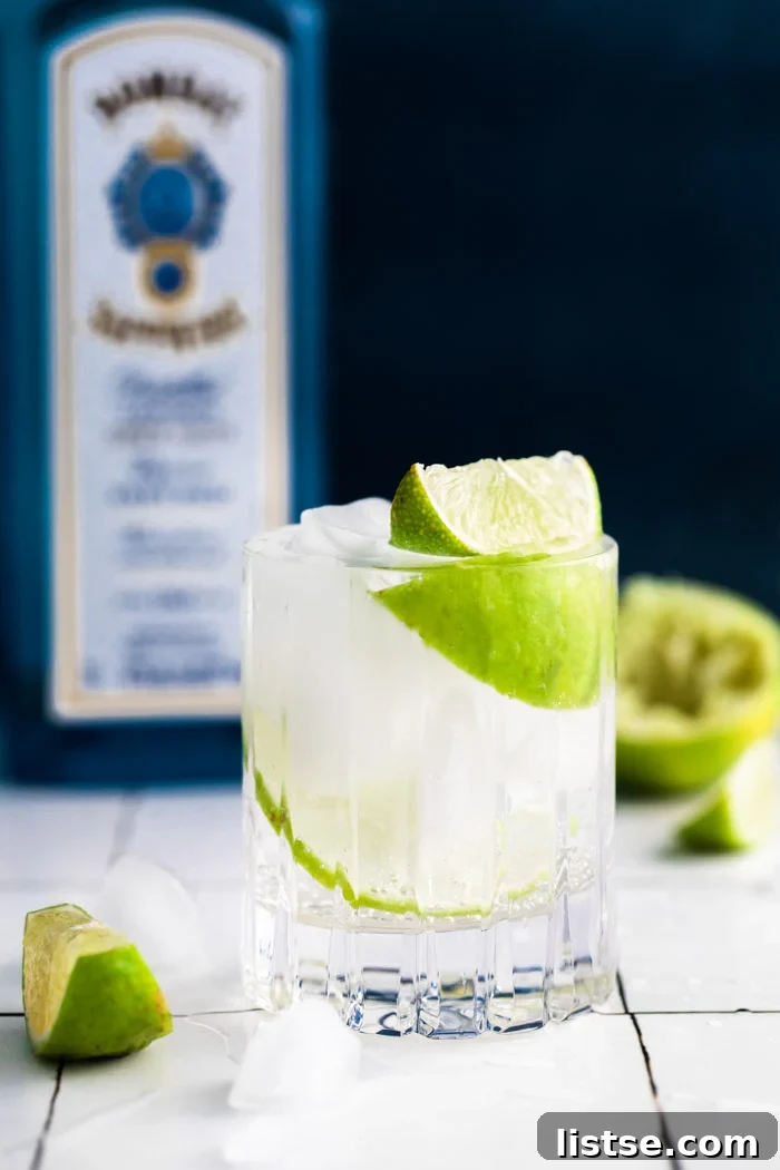 clear beveled glass filled with clear liquid (gin and tonic) plus fresh lime wedges. bombay sapphire in the background