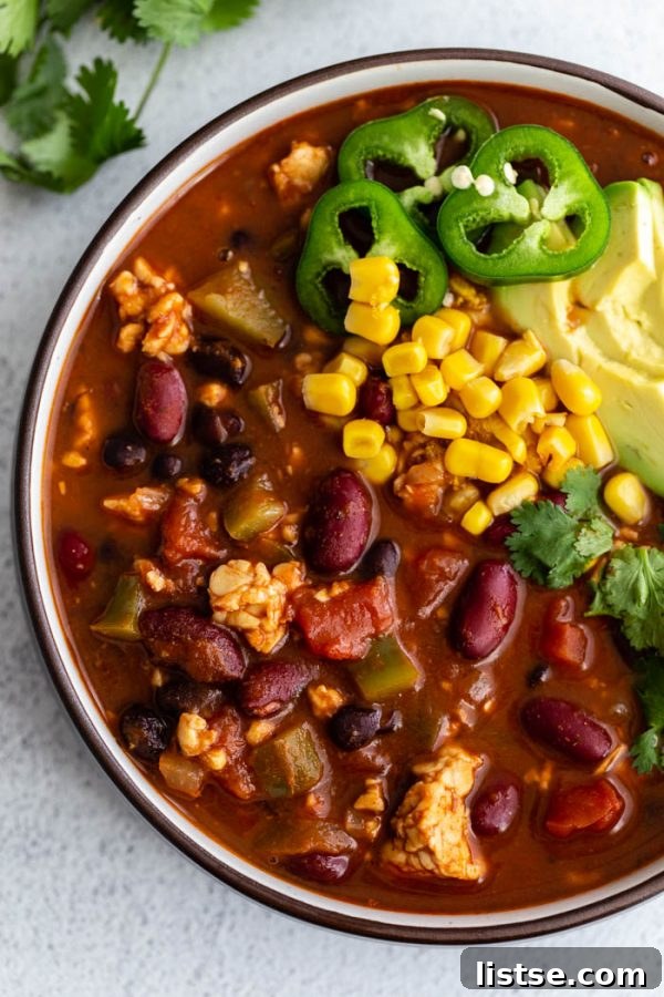 A large bowl of hearty tempeh chili, richly colored and topped with fresh corn kernels, sliced jalapeños, and creamy avocado slices, ready to be enjoyed.