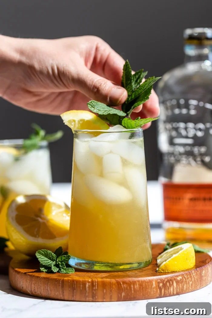 Minted Amber Elixir 2 A vibrant yellow-orange Whiskey Smash cocktail, garnished with a lemon slice and fresh mint, sits on a rustic wooden board. A bottle of bourbon is softly blurred in the background, with a hand gently placing a mint sprig into the drink.