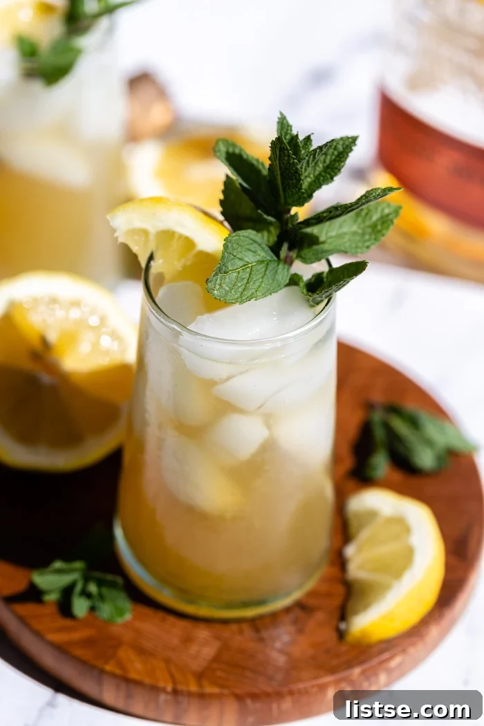 Minted Amber Elixir 3 A glistening yellow-orange Whiskey Smash cocktail, garnished with a vibrant lemon slice and fresh mint, rests on a wooden board. A bourbon bottle is visible in the soft background, adding to the rustic charm of the setup.