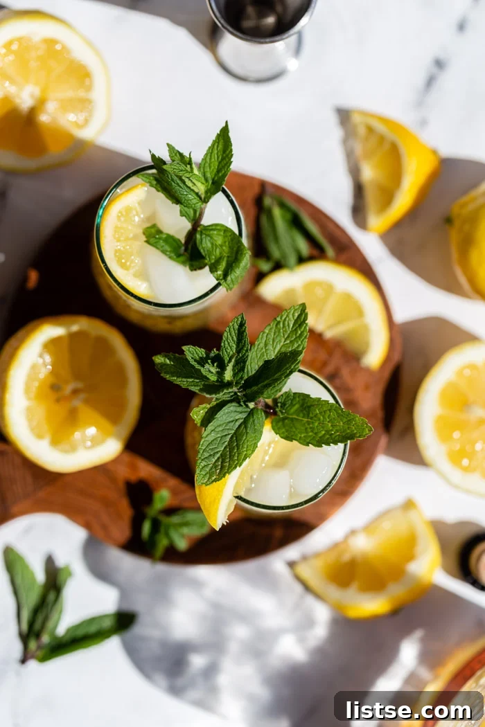 Minted Amber Elixir 4 A top-down view of two beautifully crafted whiskey cocktails, garnished with fresh lemon slices and mint sprigs, artfully arranged on a round wooden board. The vibrant colors and fresh garnishes highlight the refreshing nature of the drinks.