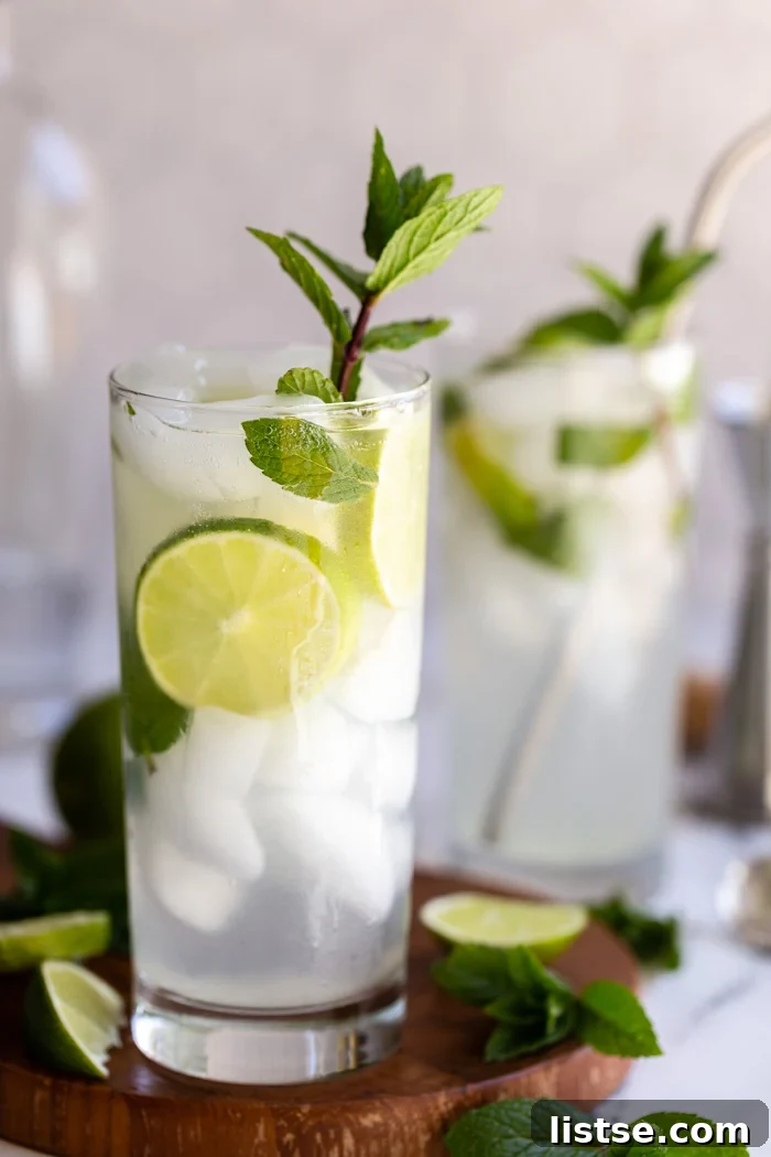 Two elegant mojitos, beautifully presented in tall glasses, complete with fresh lime and mint garnishes. The drinks are placed on a charming round wooden board against a backdrop of light, textured tiles.