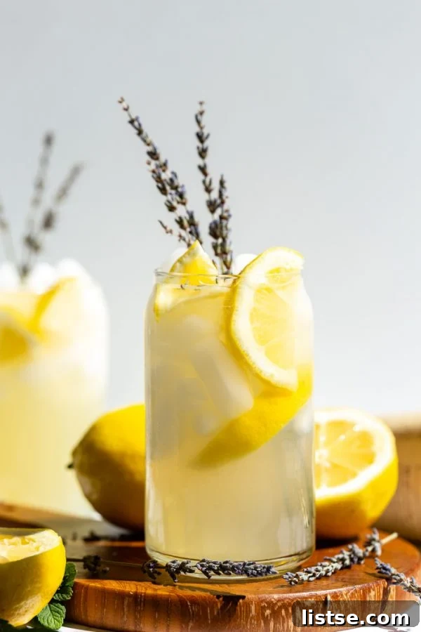 glass of lemonade with slices of lemon and lavender in it. sitting on a wood board with a white background