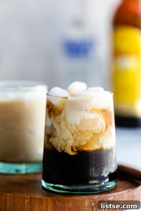 Crafting the Perfect White Russian 4 small cup of a white russian: black liquid on ice with cream floating at the top of the glass. on a wood board with white background