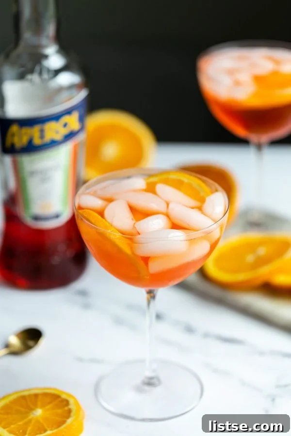 Timeless Aperol Spritz 4 An Aperol Spritz cocktail in a elegant wine glass, adorned with an orange slice, resting on a white marble surface with a blurred background of oranges and an Aperol bottle.