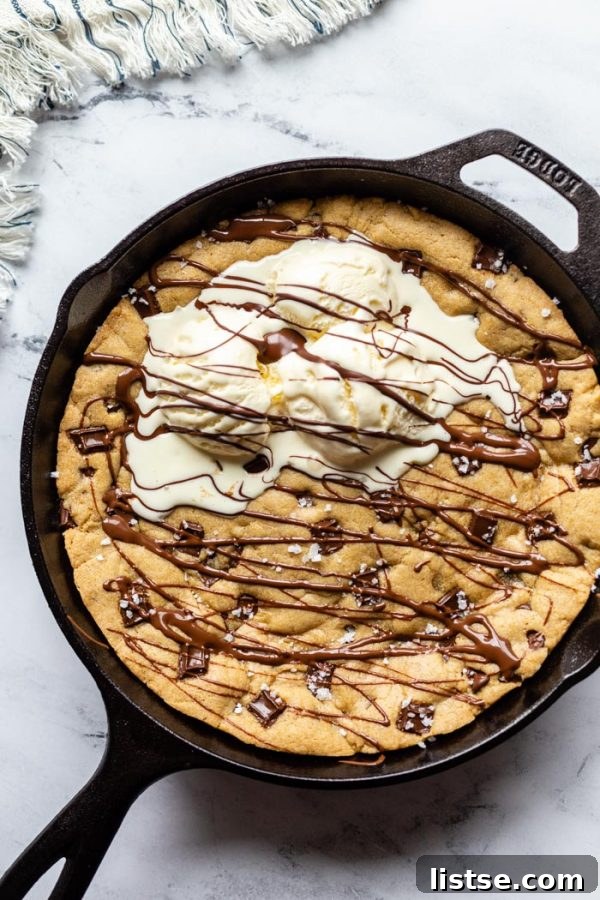 top down shot of black cast iron skillet with a skillet cookie in in covered in chocolate chunks, sea salt, and topped with 3 scoops of ice cream and drizzled with chocolate syrup. On a marble background with a stripped towel in the corner